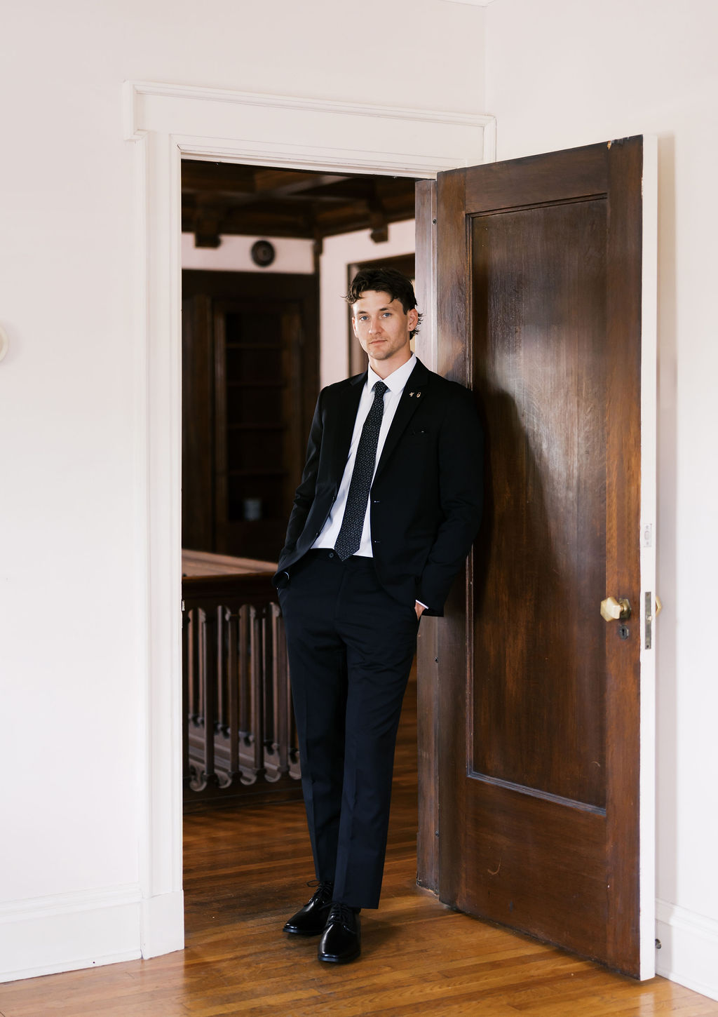 Person in a black suit stands in a wooden doorway inside a bright, historic-style interior.