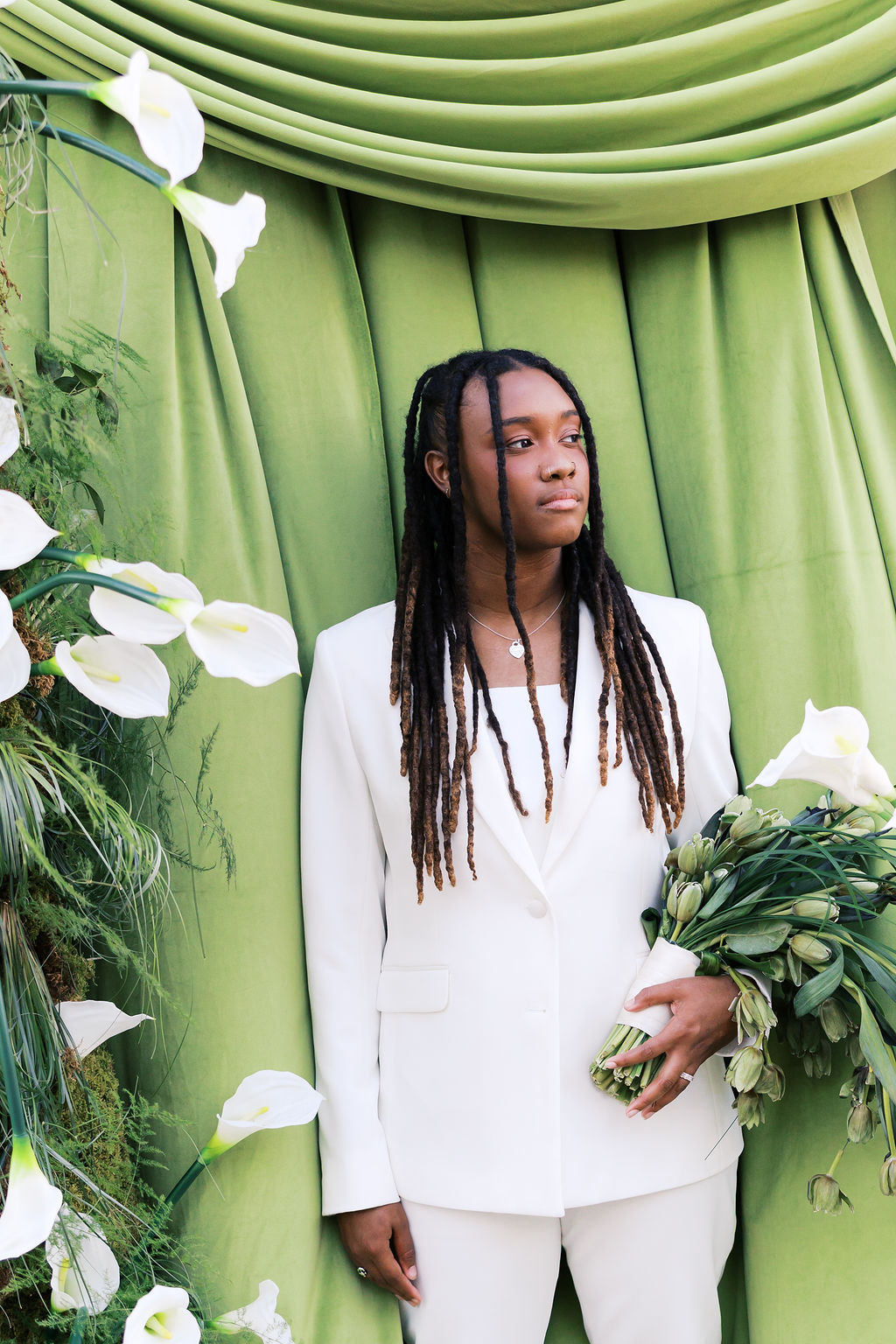 Person in a white suit holds a green floral bouquet in front of draped green fabric and white calla lilies.