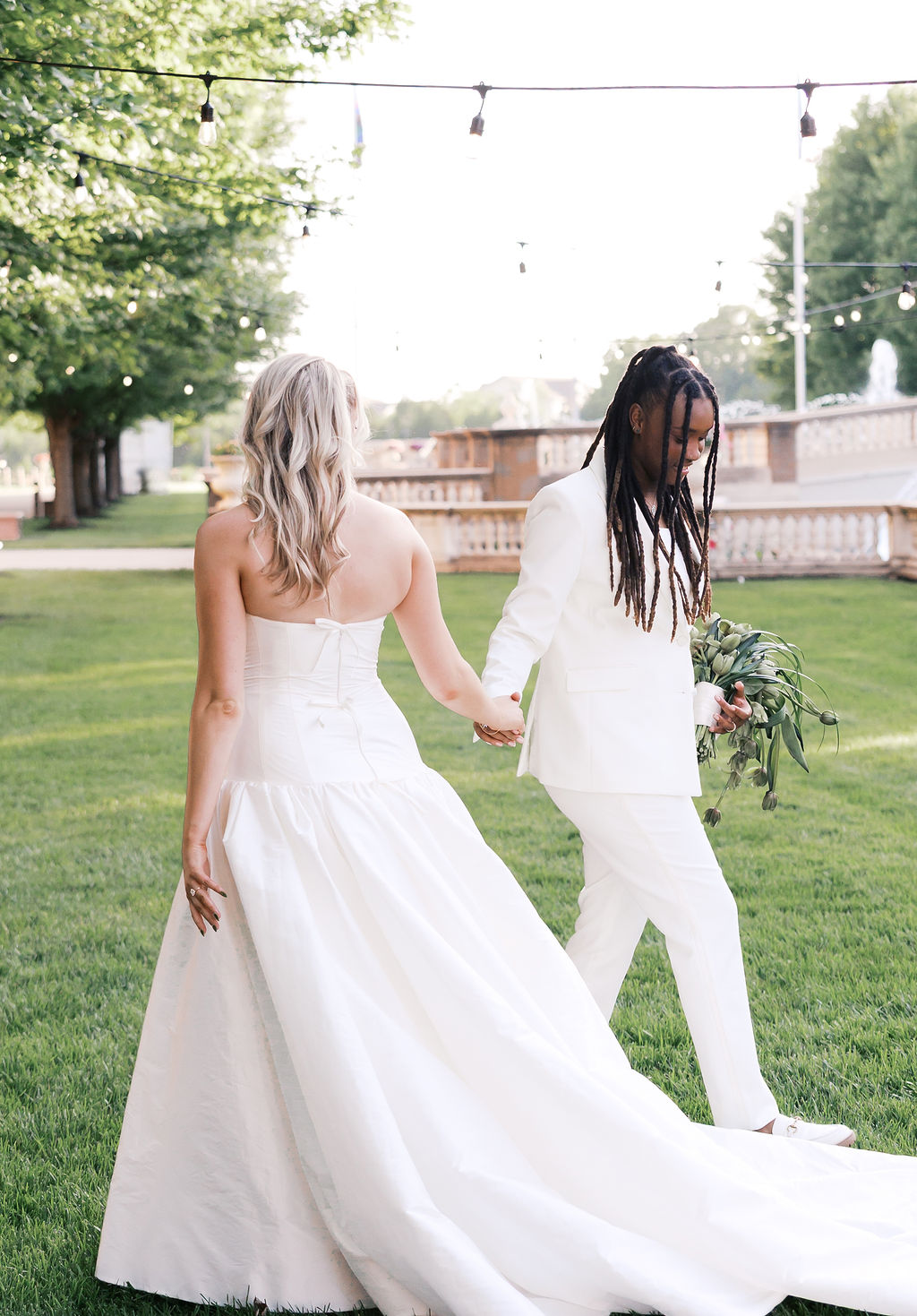Two people in white wedding attire hold hands while walking across a sunlit lawn beneath string lights.
