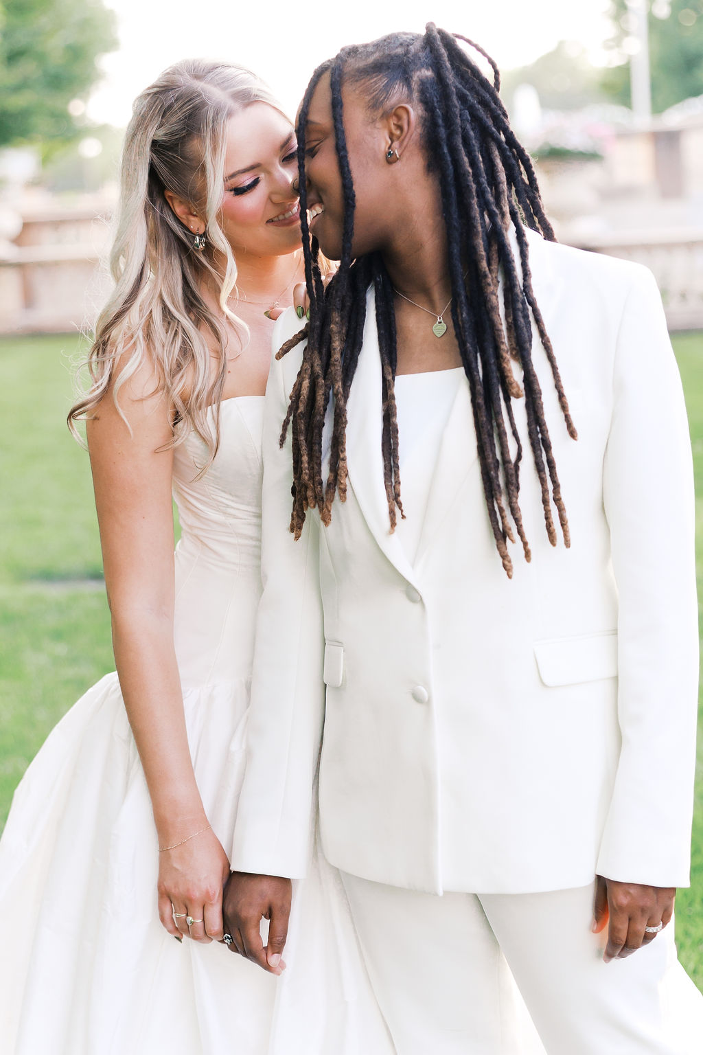 Two people in white wedding attire stand closely together during an outdoor portrait on a green lawn.