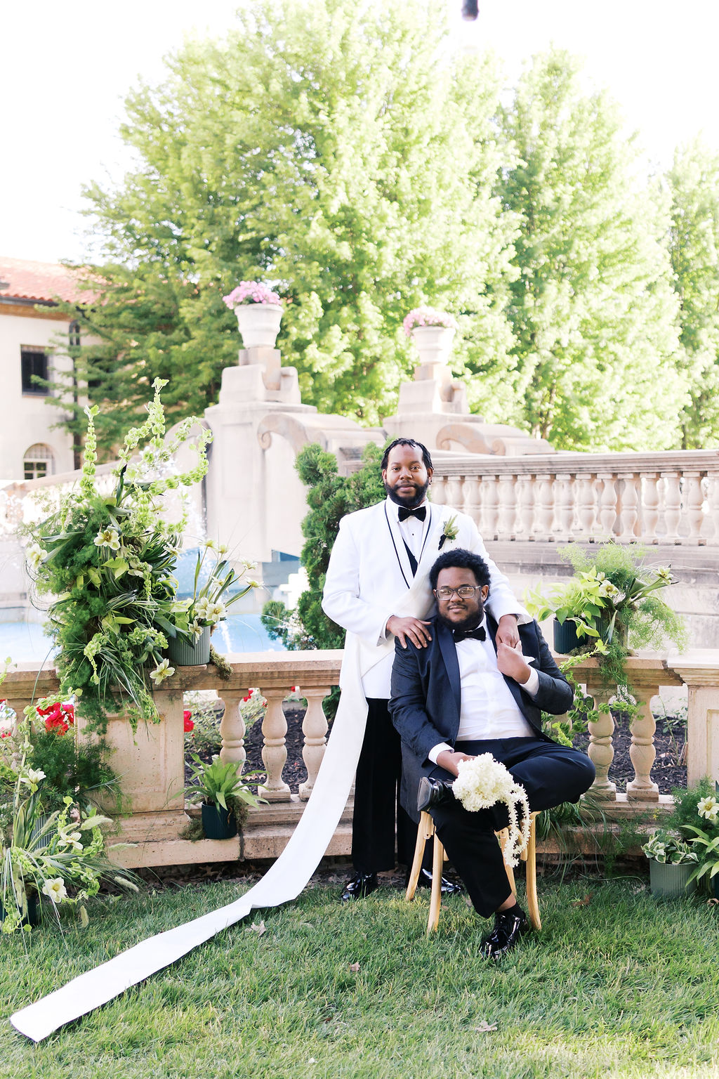 Two people in formal wedding attire pose in a garden surrounded by lush greenery and floral arrangements.