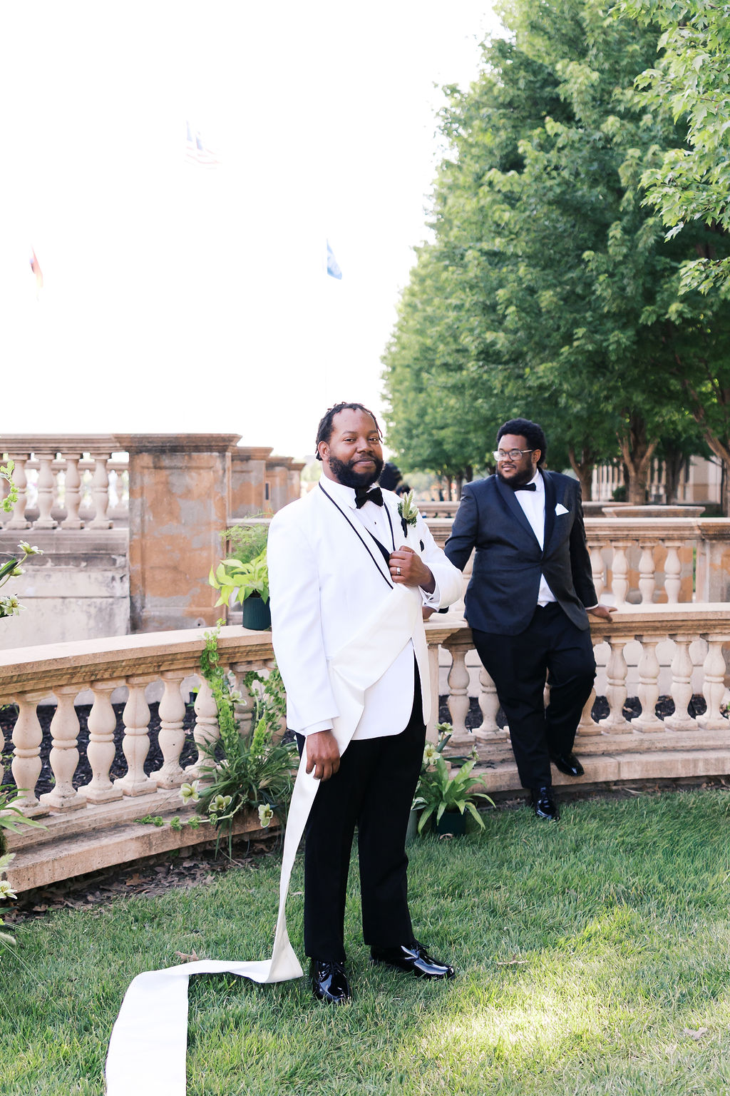 Person in a white tuxedo poses in a formal garden while another person in a dark tuxedo stands behind them.
