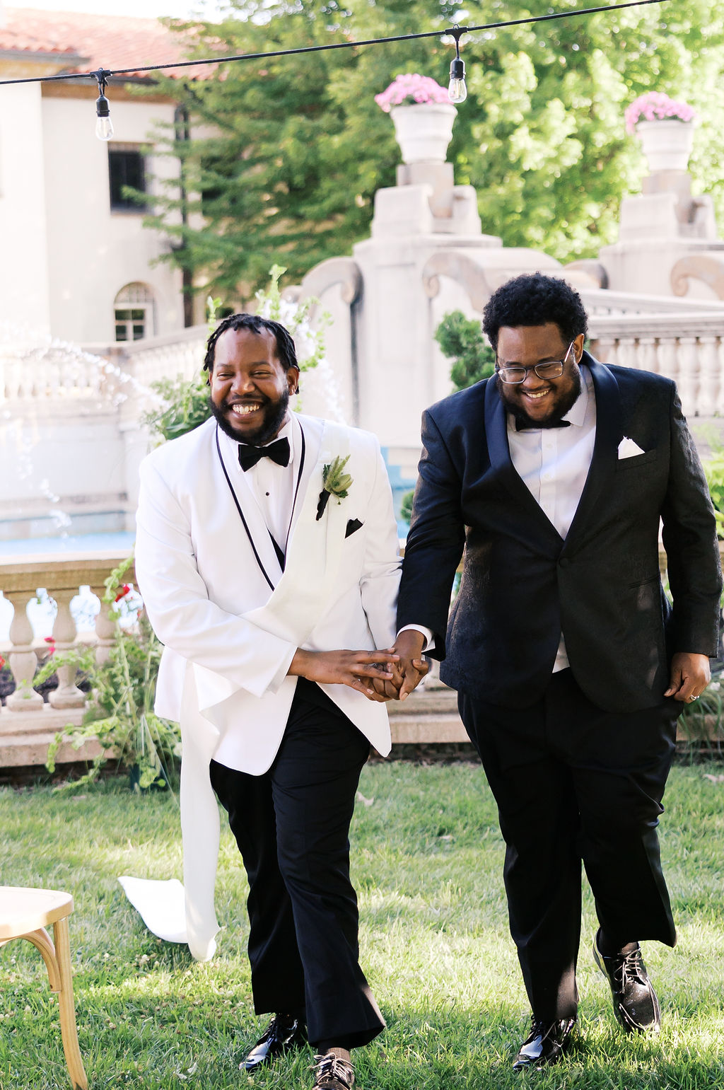 Two people in black-and-white tuxedos walk hand in hand through a sunlit garden wedding setting.