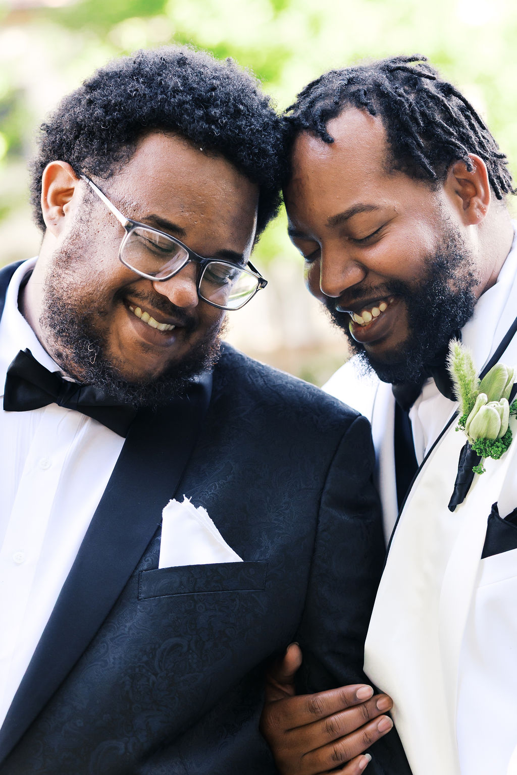Close-up portrait of two people in tuxedos smiling together outdoors, one wearing a white jacket and boutonniere.