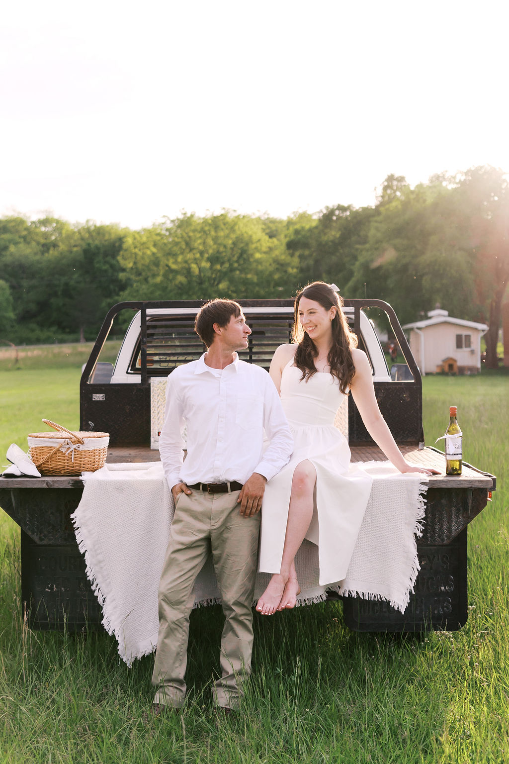 Couple in white outfits sitting on the back of a pickup truck during a sunset picnic engagement session.