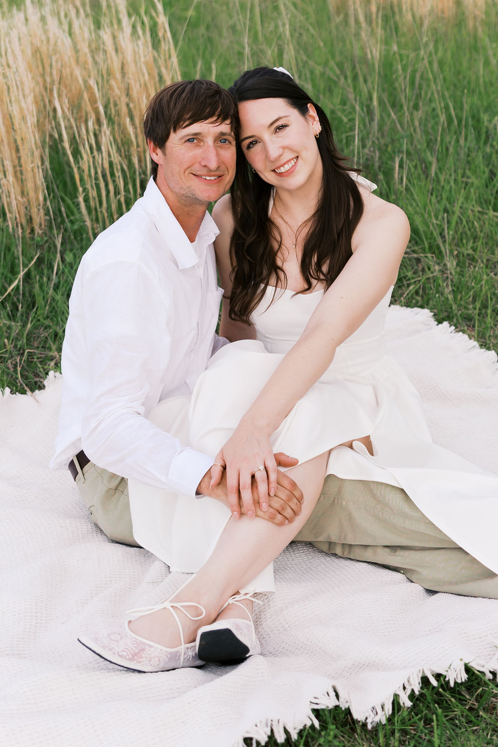 Couple sitting together on a white picnic blanket in tall grass during their engagement photos.