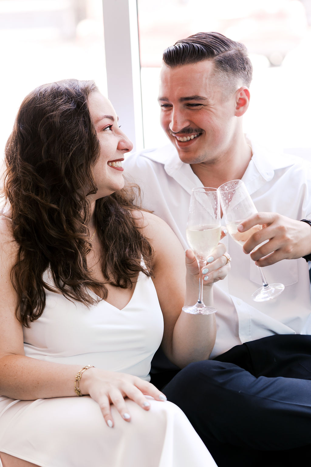 Couple smiling at each other while raising champagne glasses during their engagement session.