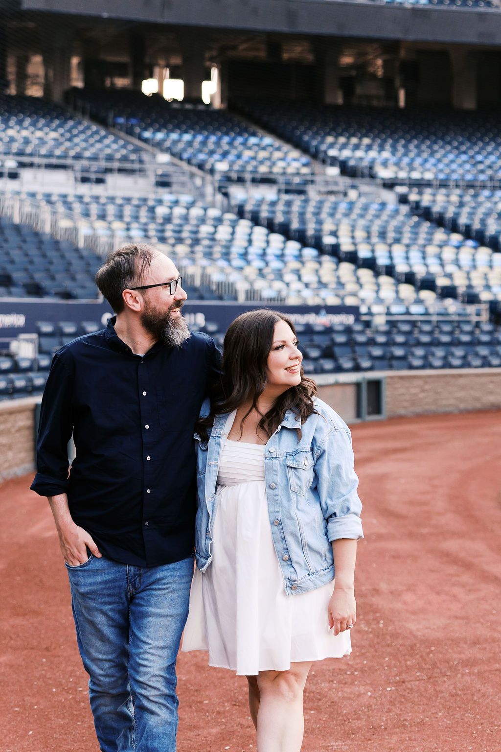 Couple standing together near the baseball field with rows of stadium seats behind them.