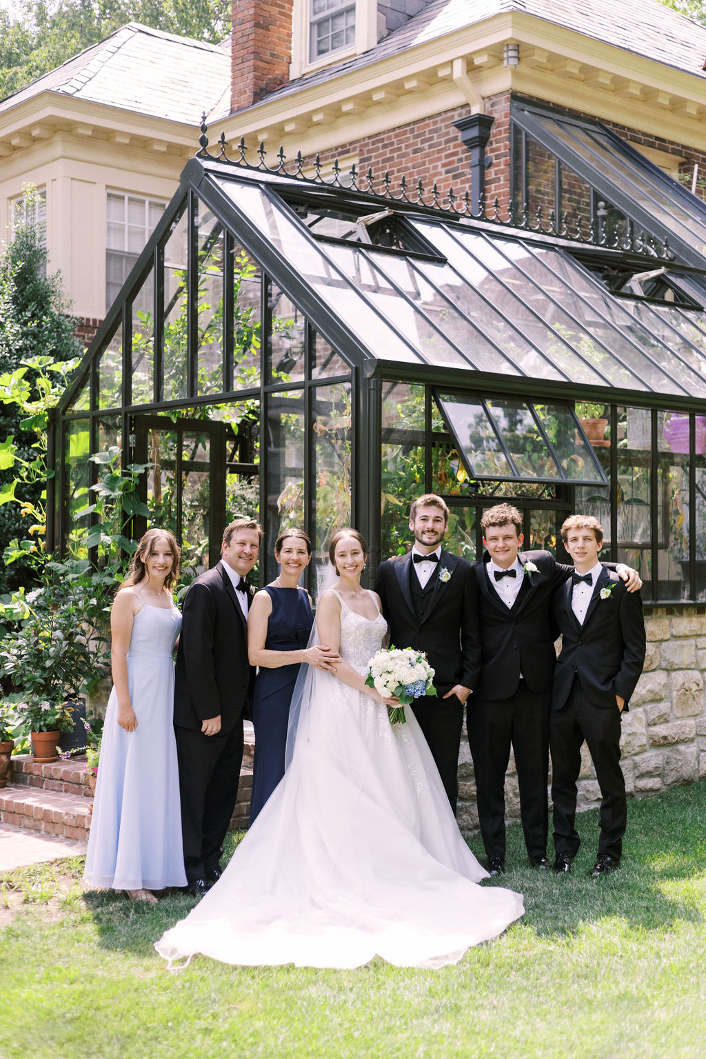 Bride and groom gather with family members for a relaxed wedding family portrait beside a greenhouse.