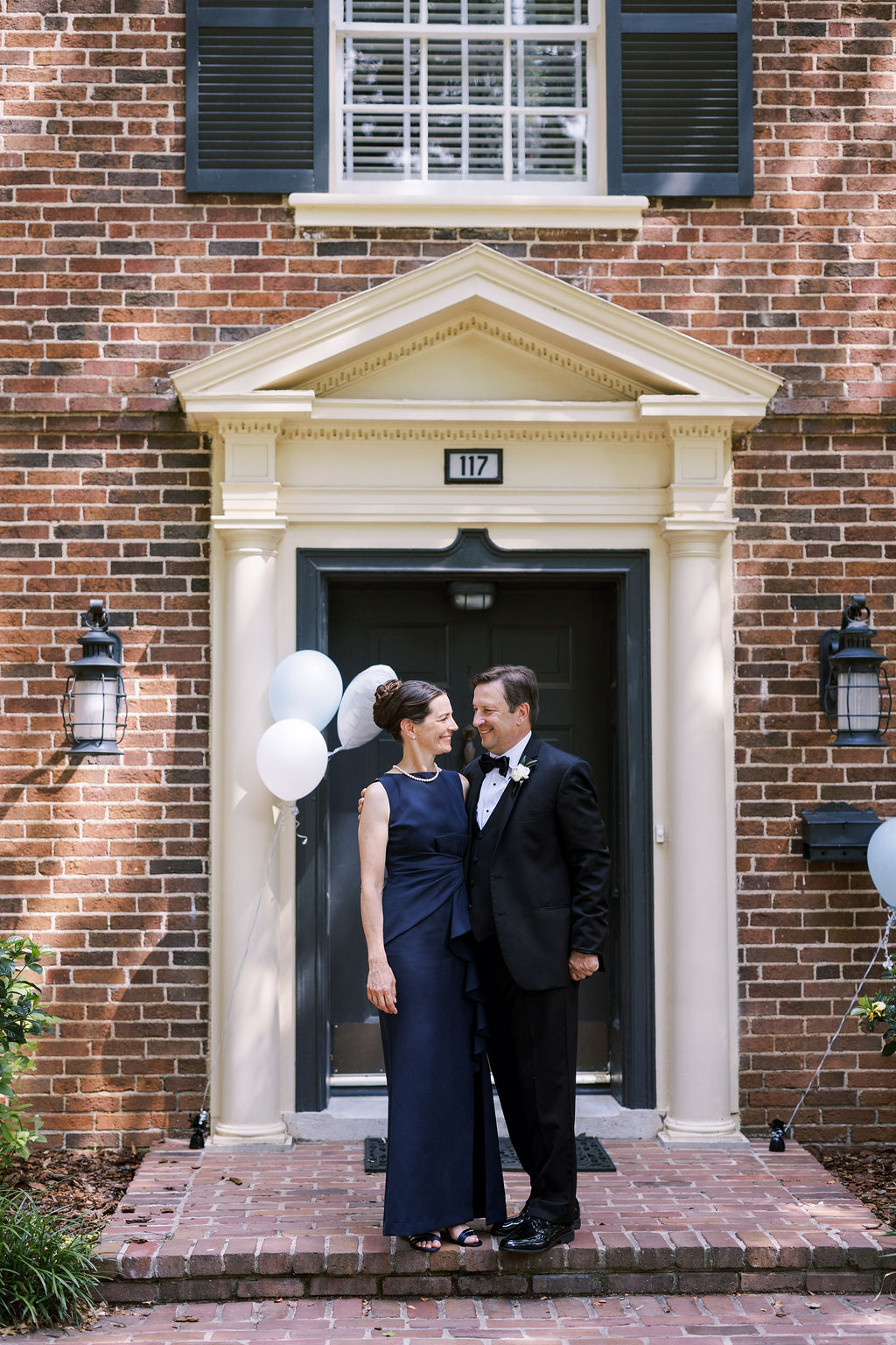 Wedding guests in formal attire pose together outside a brick home decorated with white balloons.