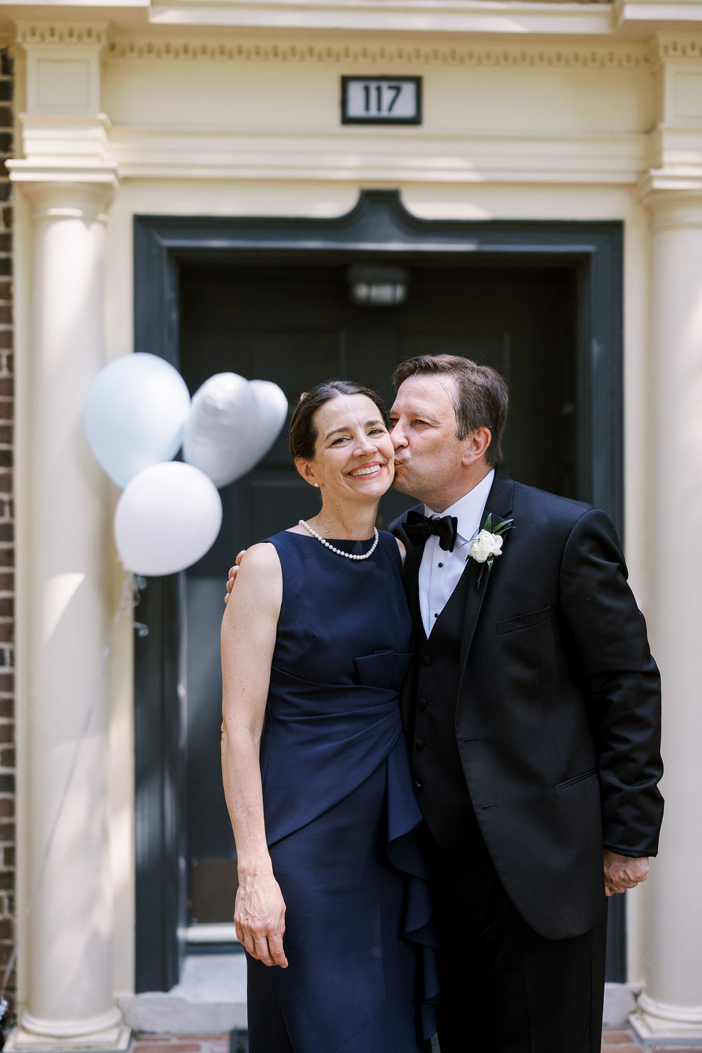 Man in a tuxedo kisses a woman in a navy dress on the cheek during a candid wedding family portrait.