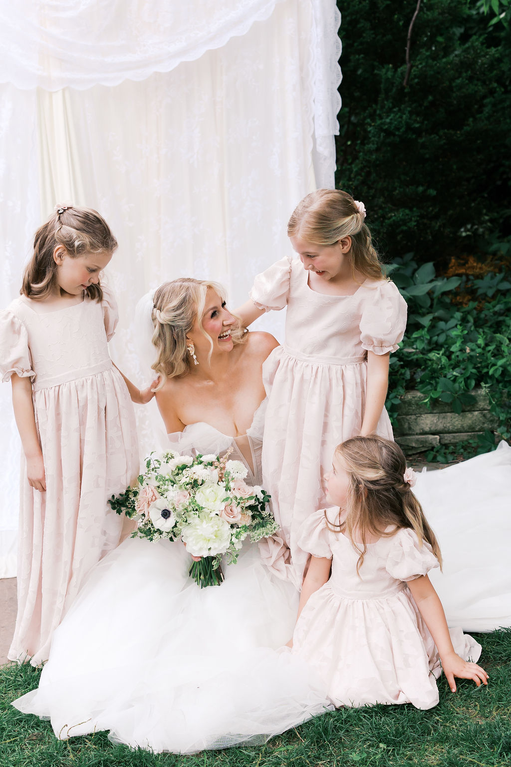 Bride sits with three flower girls in pale pink dresses during outdoor wedding family portraits.