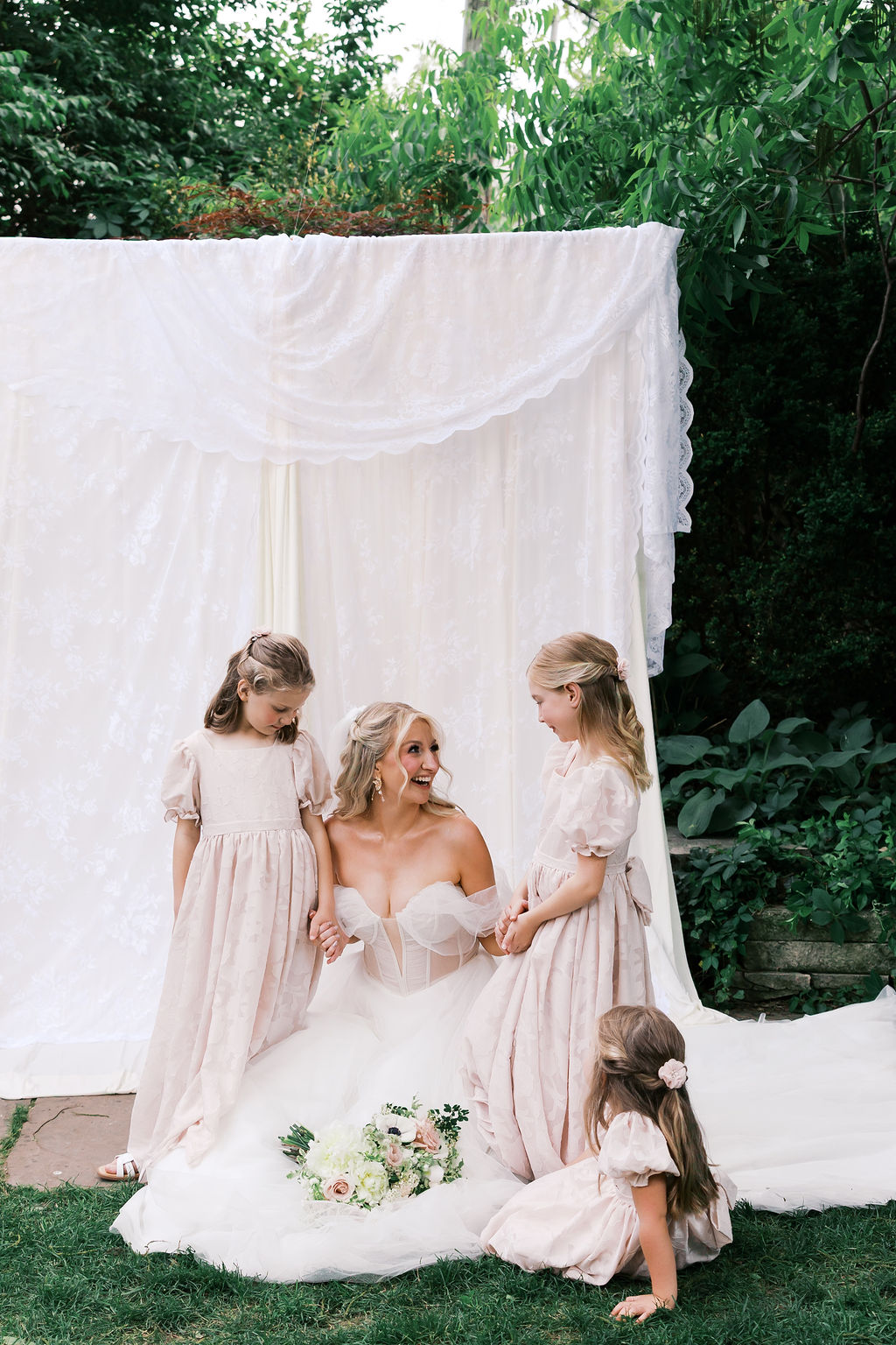 Bride talks with three flower girls beside a white fabric backdrop during relaxed wedding portraits.