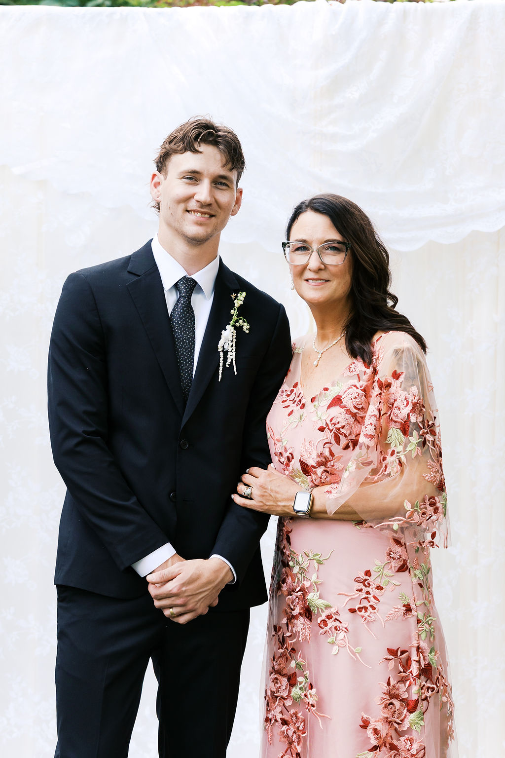 Man in a black suit poses with a woman in a floral pink dress against a soft white wedding backdrop.