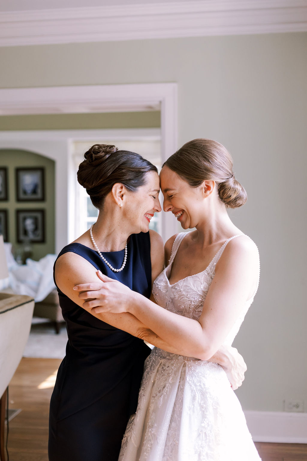 Bride and a woman in a navy dress embrace and laugh together during a candid indoor wedding moment.