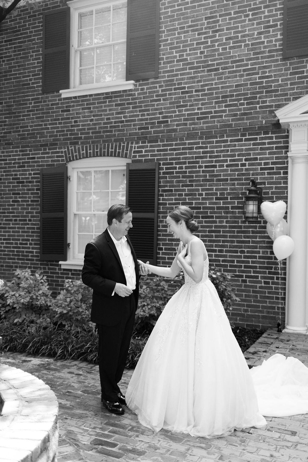 Black-and-white portrait of a bride and tuxedoed man laughing together outside a brick home.