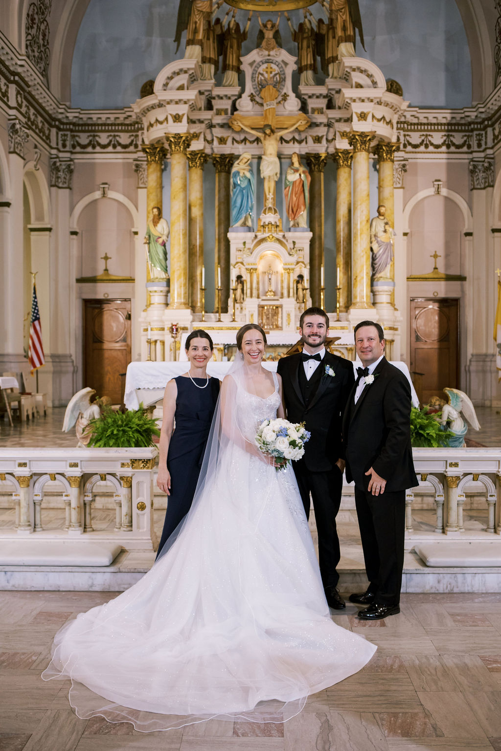 Bride and groom pose with two family members in front of an ornate church altar after the ceremony.