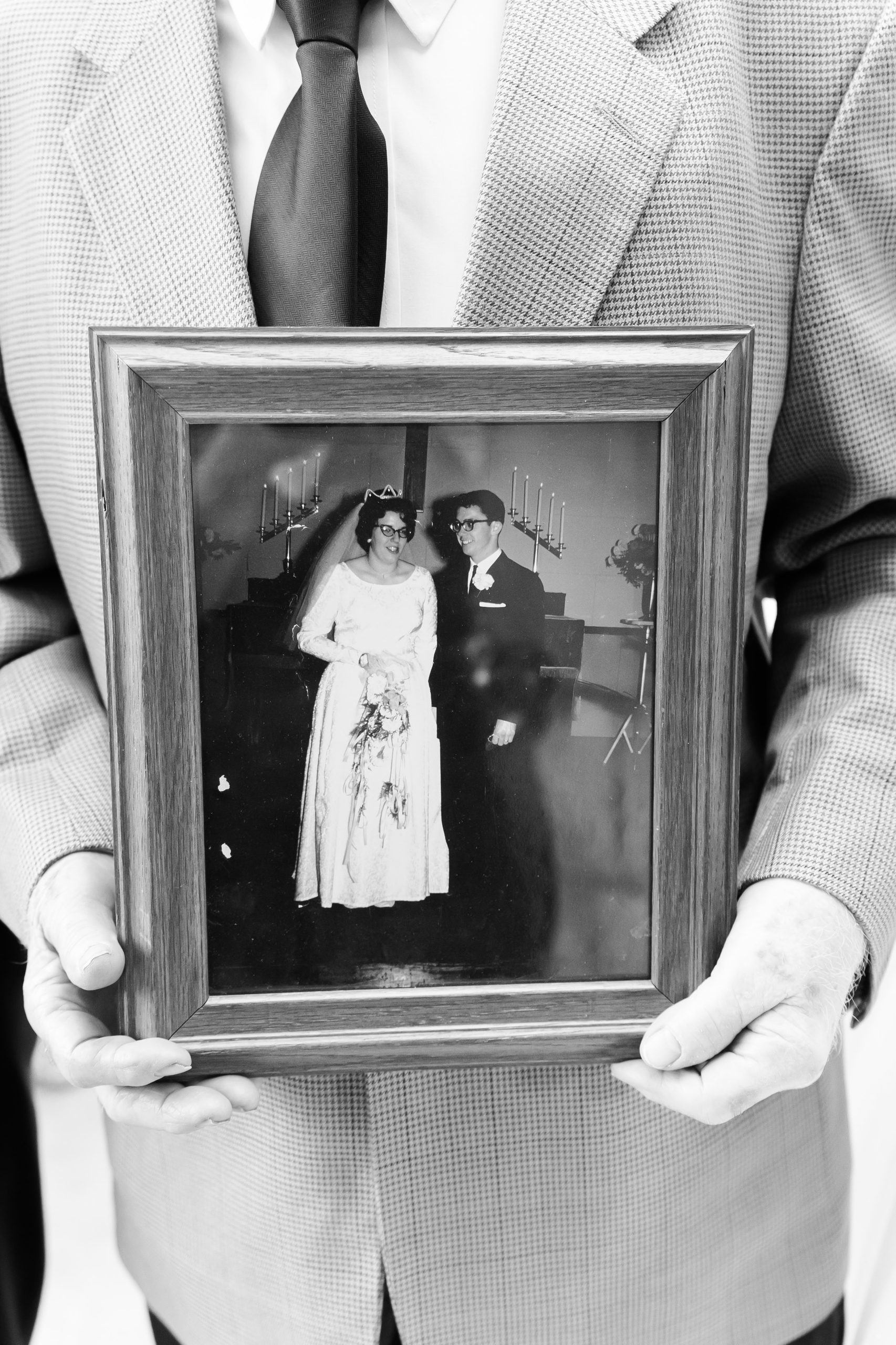 Black-and-white photograph of a person in a suit holding a framed vintage wedding portrait of a couple, preserving a meaningful family memory.