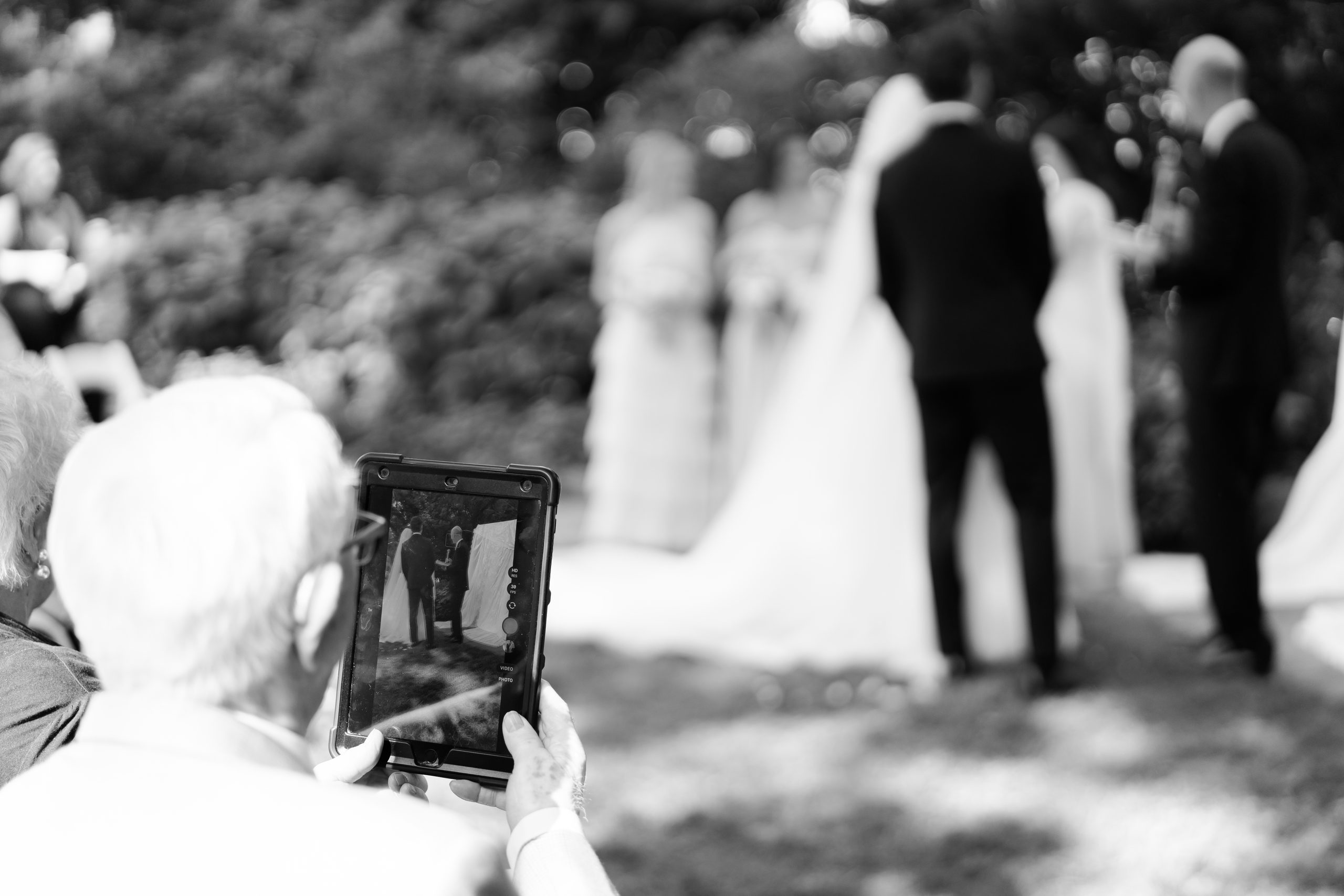 Black-and-white photograph of a wedding guest capturing an outdoor ceremony on a phone, with the couple exchanging vows in the background.