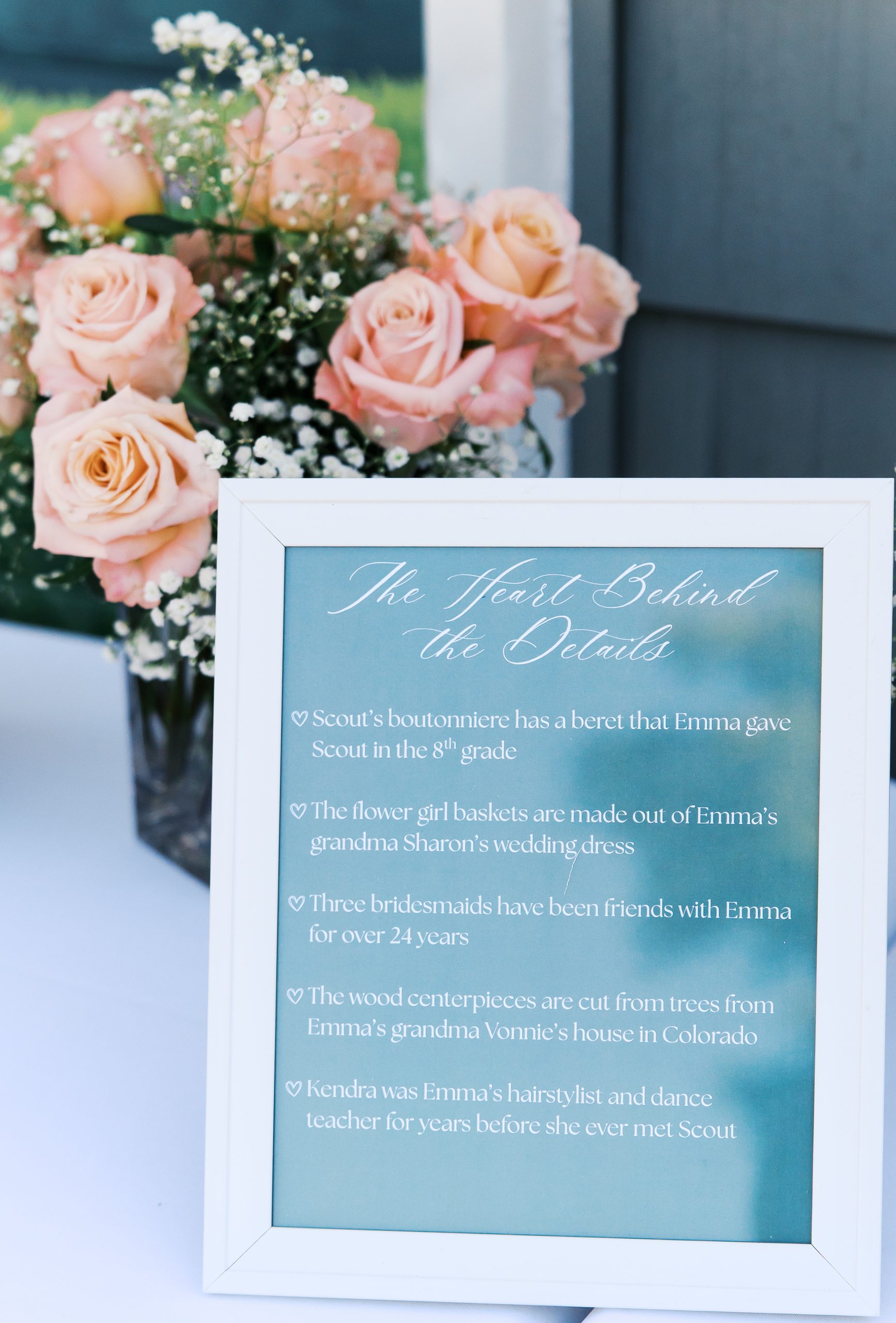 Wedding reception display featuring pink roses and a framed sign explaining meaningful personal wedding details, including family heirlooms and traditions.
