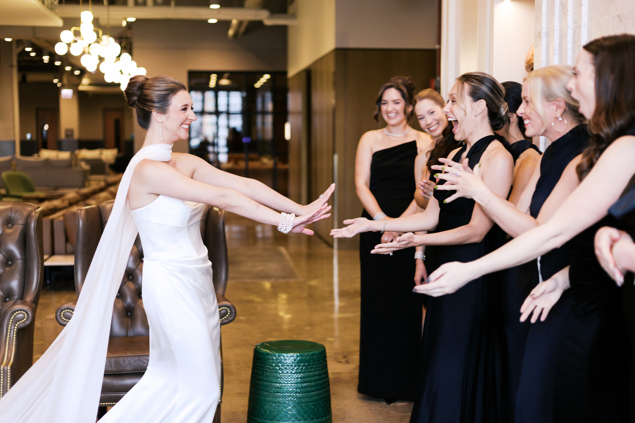A bride in a white wedding dress shares an excited moment with her wedding party, who laugh and reach toward her in matching black dresses.